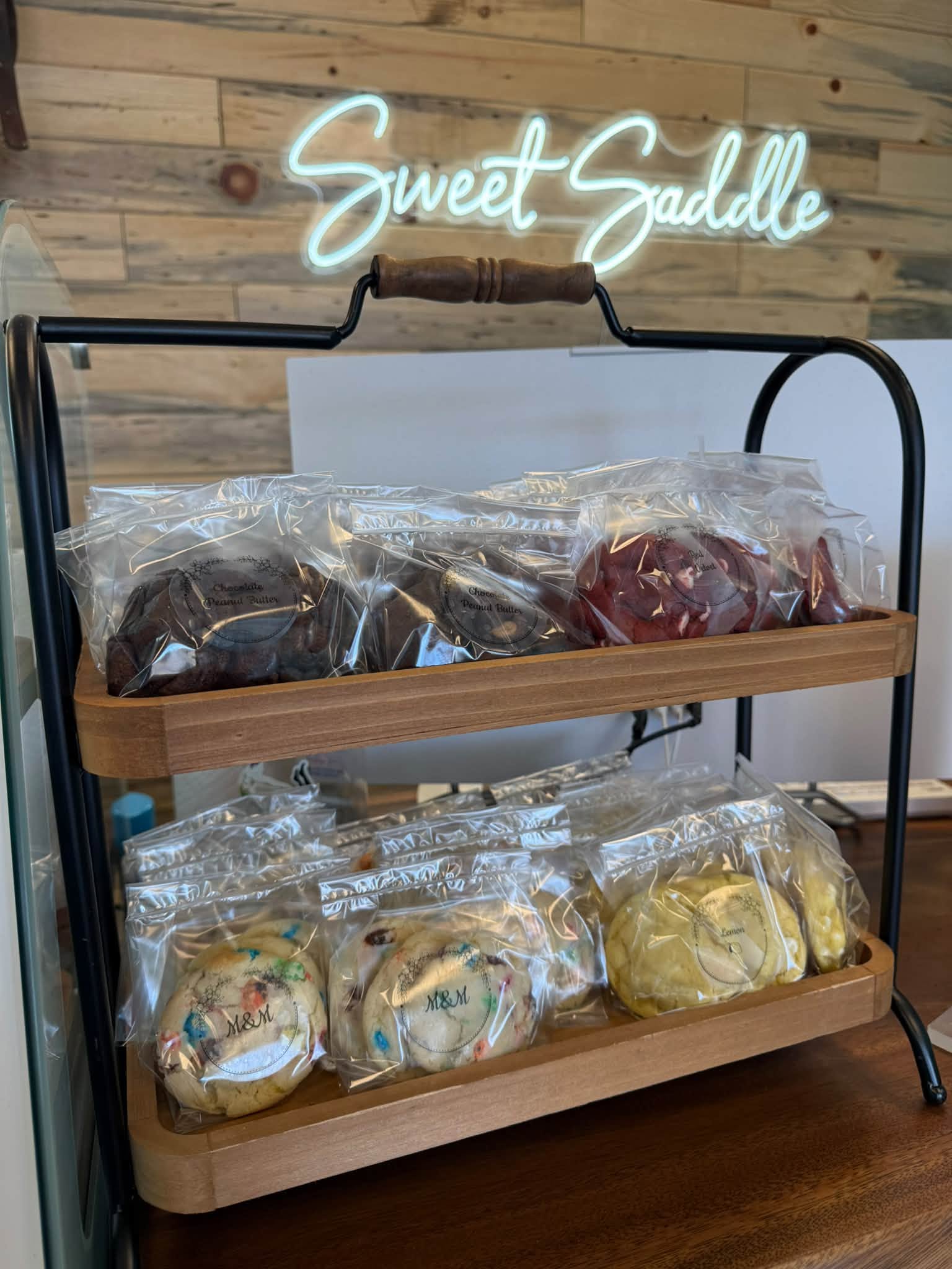 Wooden tiered tray of individually wrapped cookies beneath the Sweet Saddle neon sign