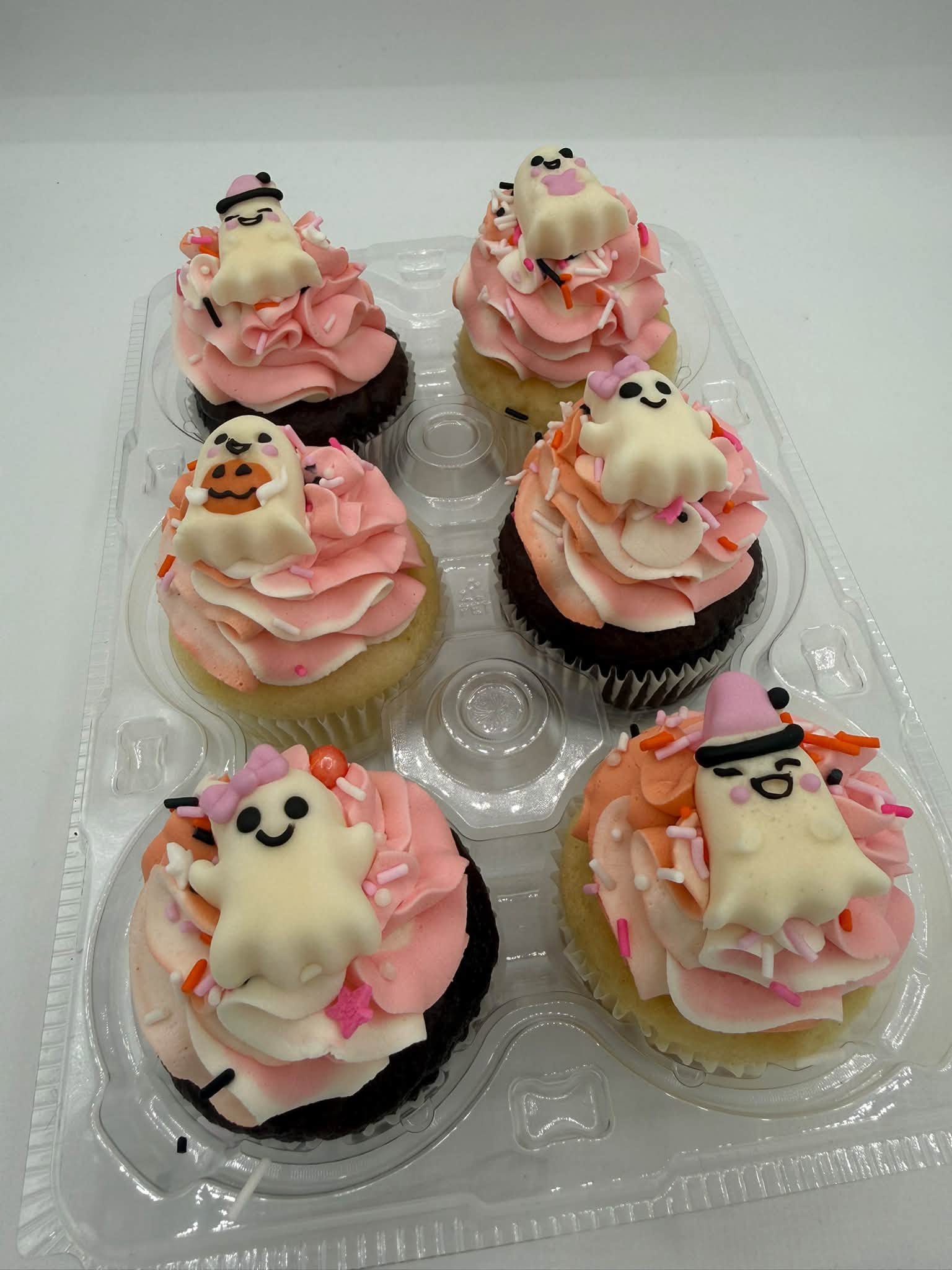 Six cupcakes in pink and orange buttercream topped with smiling ghost decorations
