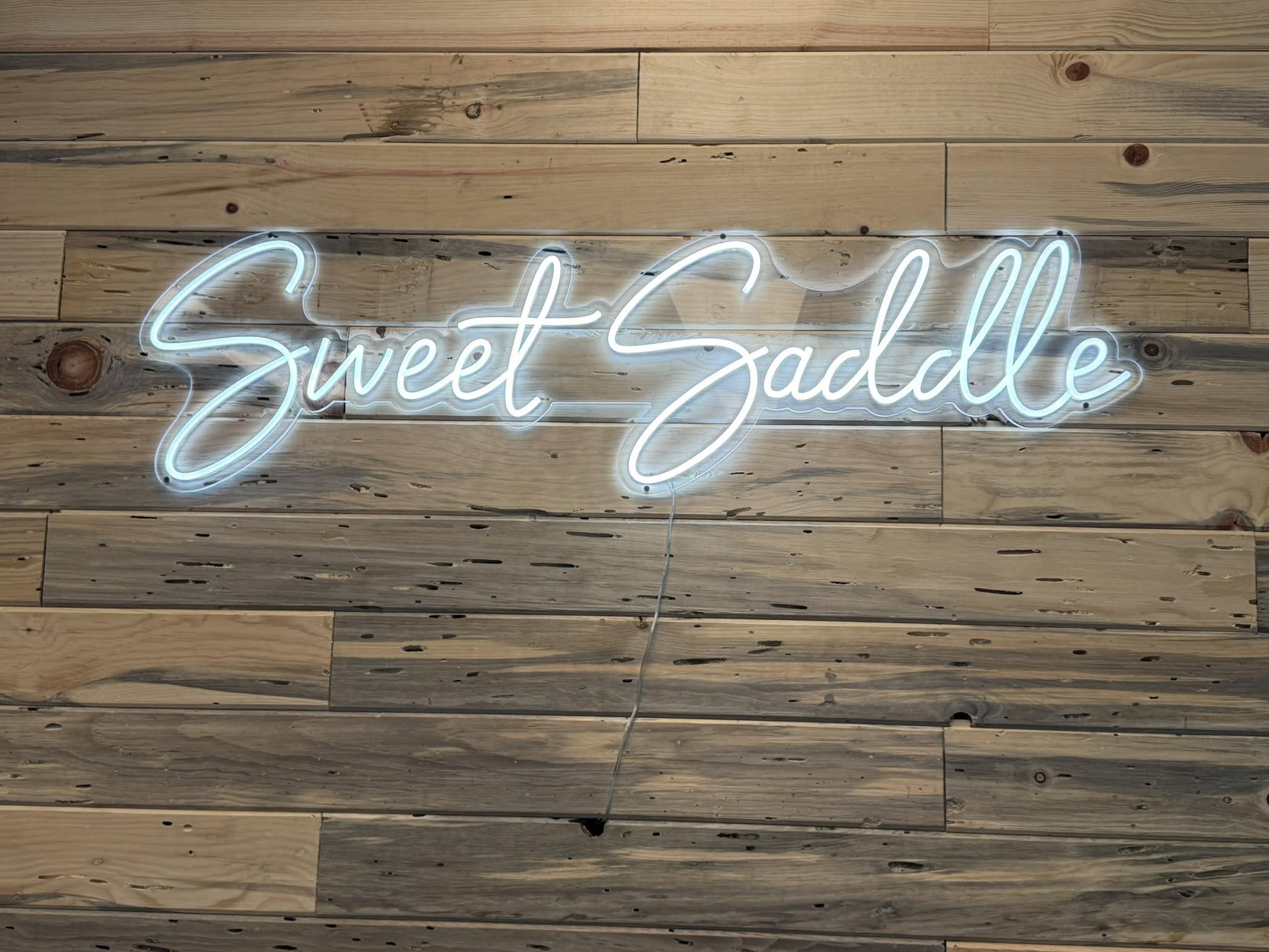 White neon Sweet Saddle script sign on a reclaimed pine plank wall