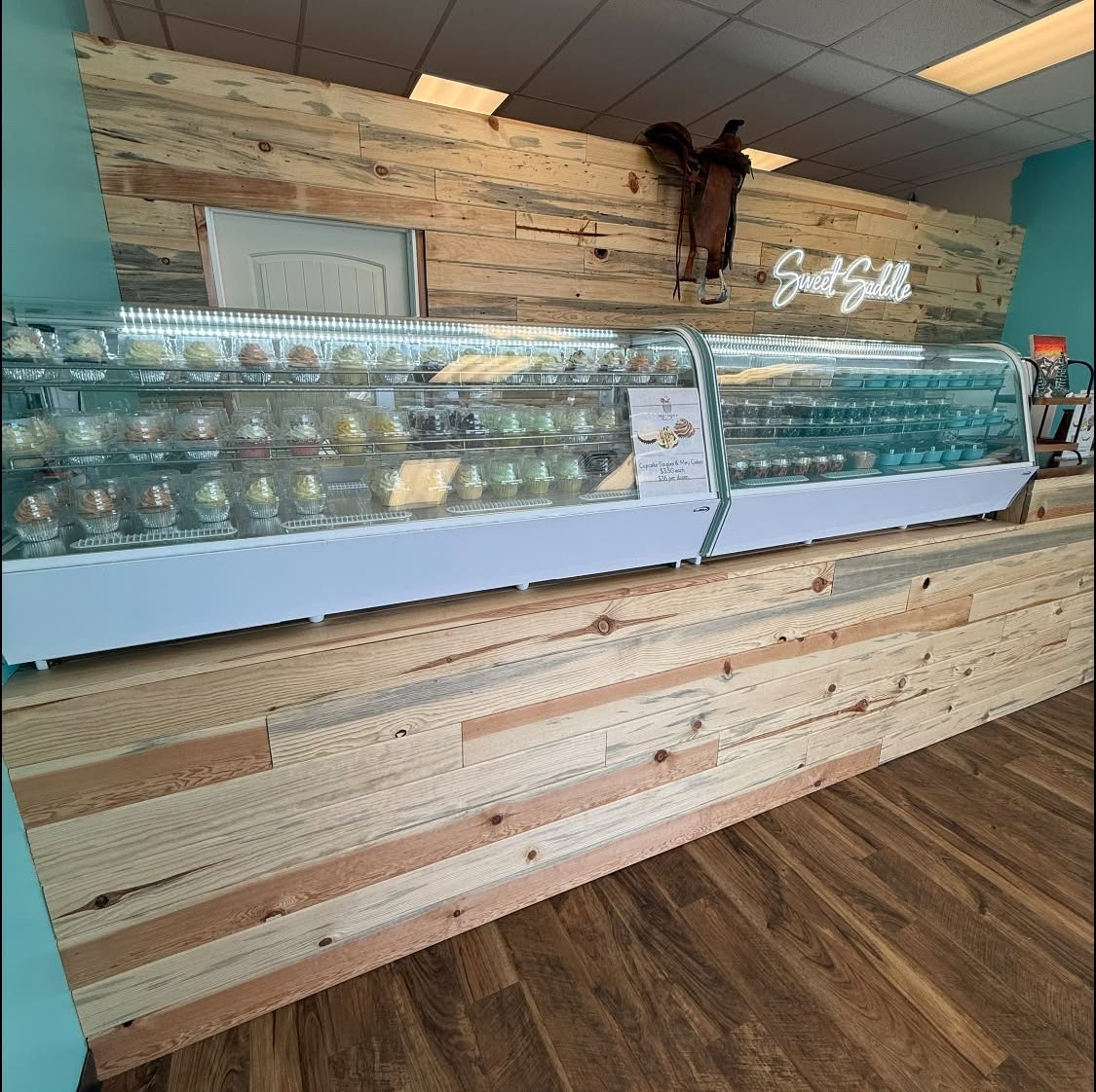 Long lit display case full of cupcakes in front of a pine wall with a saddle hung above